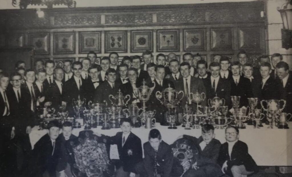 A black and white photo of the rowing club gathered around a trophy table.